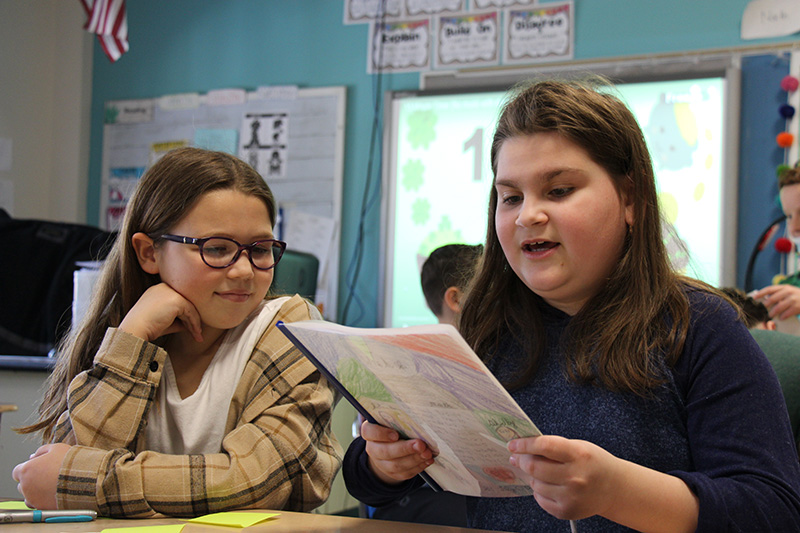 Two fifth grade girls sit together. The one on the left is wearing a beige plaid shirt and glasses, looking at the girl on the right. The girl onthe right has long dark hair and is reading a book she made.