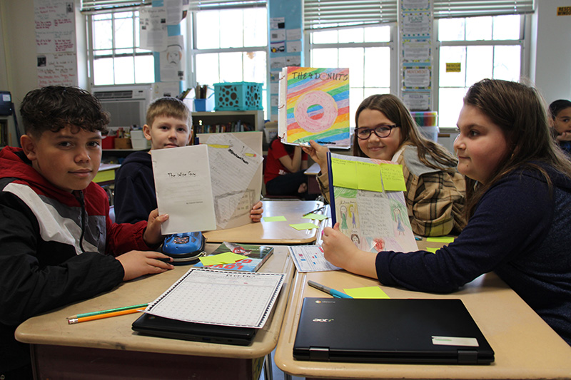 A group of four fifth grade students sit at a group of desks together, holding up the books they made. Two boys and two girls