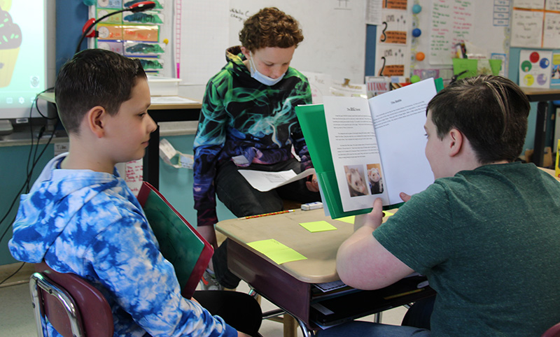 Three fifth grade boys sit at desks. One is reading a handmade book and the other two are listening.