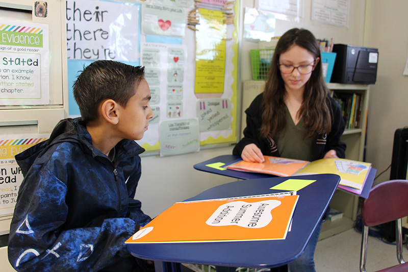 Two fifth grade students sit at desk, each with homemade books. 