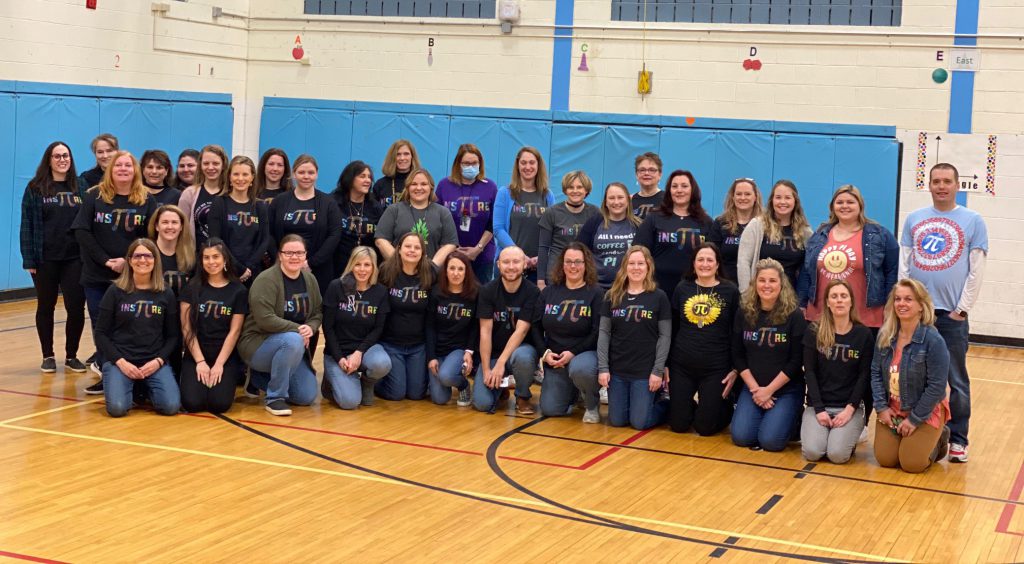 A large group of adults in a gymnasium. They all have shirts on that say something about the math figure pi. Many say inspire with the pi replaced with the Greek symbol for it.