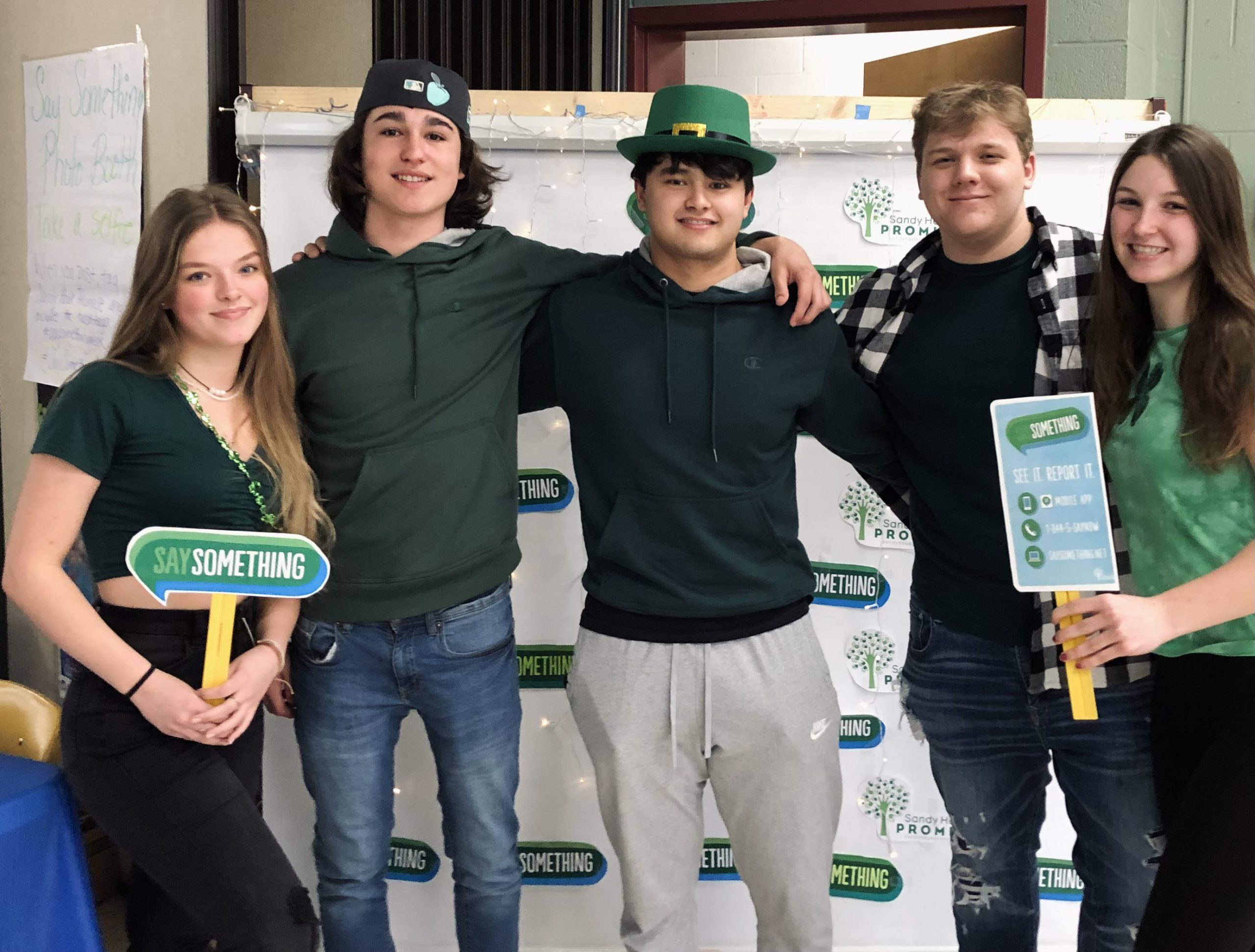 Five high school students stand in front of a banner, white with green messages saying Say Something. The girls on the ends are  holding Say Something signs. The three boys in the center have all locked arms. The boy in the center is wearing a green hat.