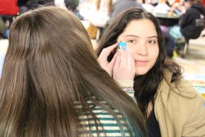 A high school girl with long dark hair paints another high school girs face. She too has long dark hair.