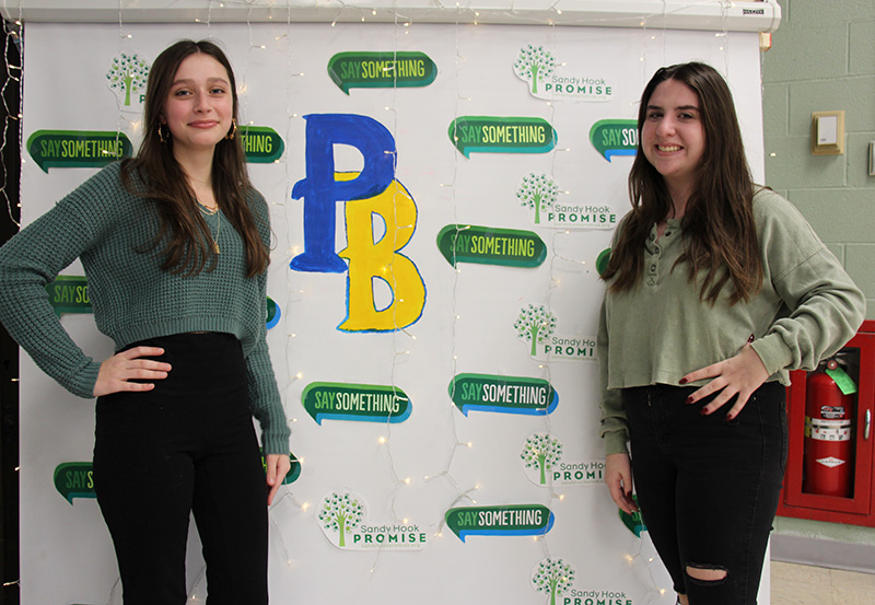 Two high school girls stand on either side of a banner,  with a white background and green talk bubbles that say Say Something. There is a big PB in the center, in blue and gold.