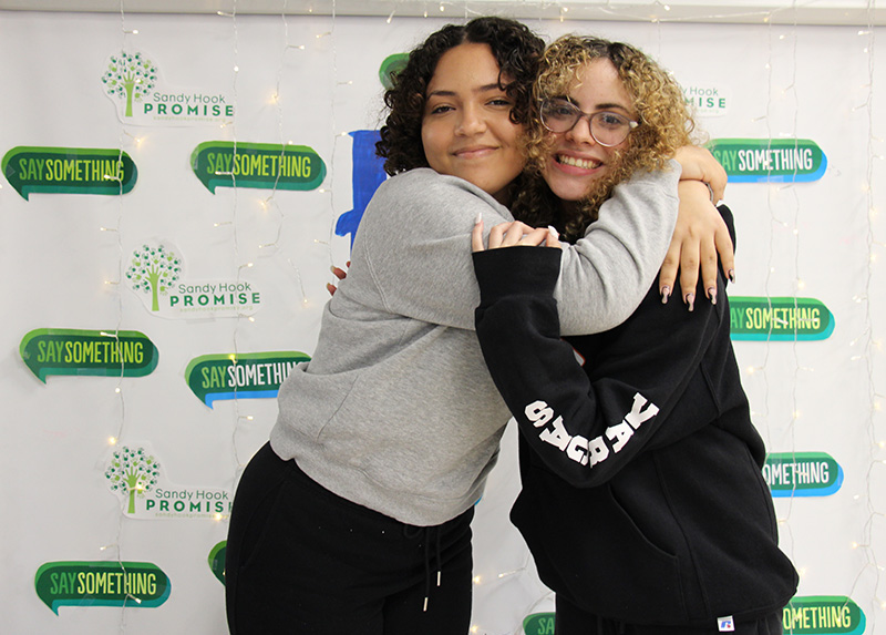 Two high school girls hug each other in front of  a banner with a white background and green talk bubbles that say Say Something. There is a big PB in the center, in blue and gold.