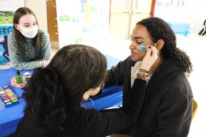 A high school girl uses a q-tip to paint another student's face.