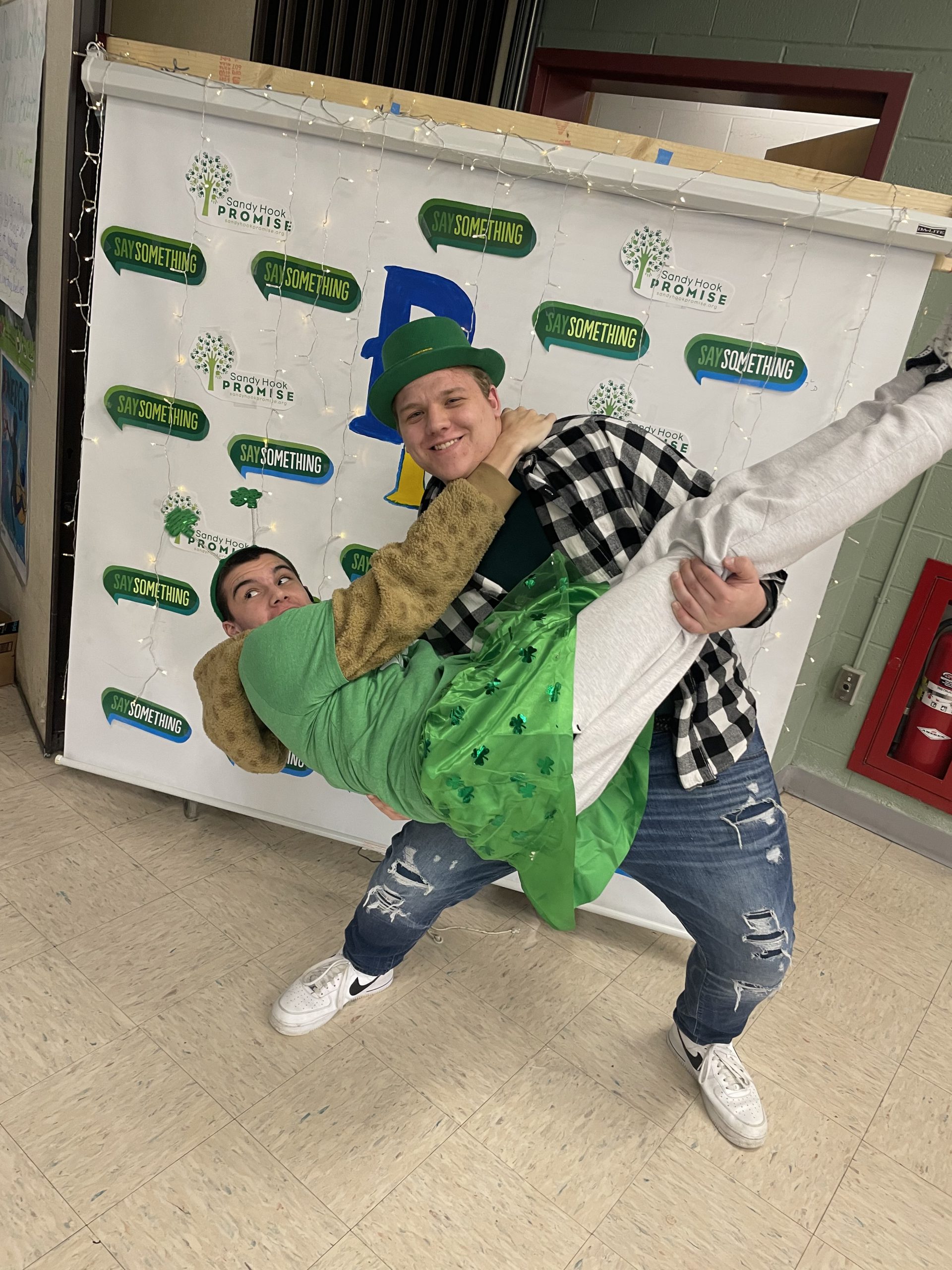 Two high school students clown around in front of a banner with Say Something all over it. The one boy, wearing a green hat and a black and white checked shirt smiles as he holds another high school boy , wearing white pants and a green shirt and tutu.