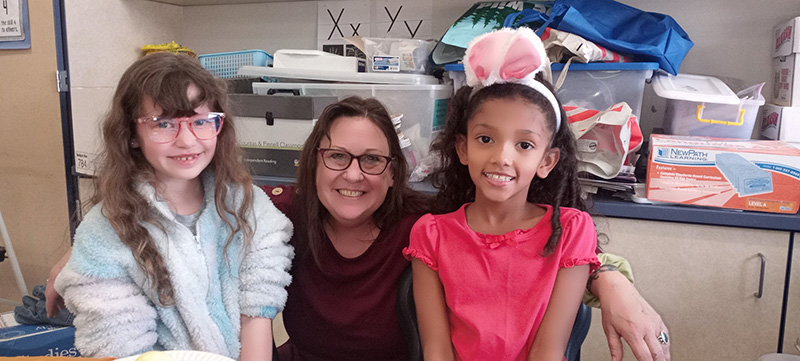 A woman squats between two girls sitting on chairs. They are second grade students. Girl on the left is wearing a blue fuzzy jacket, has long brown hair and is wearing red glasses. The girl on the right has on a pink shirt, long brown hair and is wearing a white headband with pink bunny ears. The woman in the center has long dark hair, and is wearing a maroon shirt and glasses. All are smiling.
