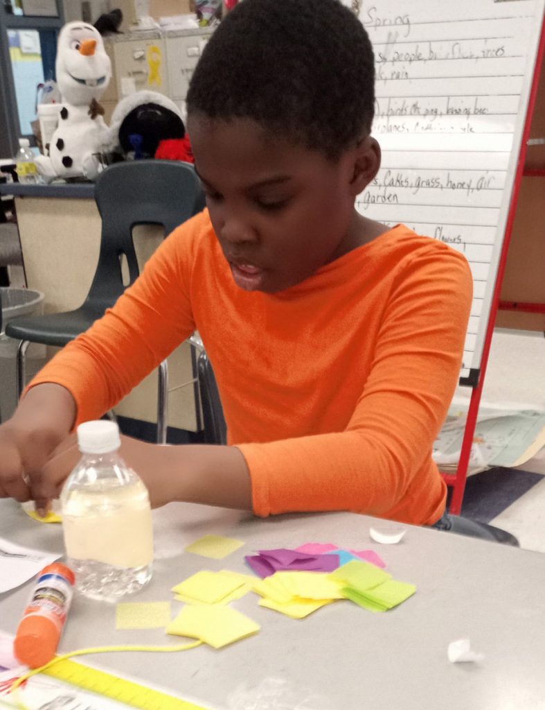 A boy with short dark hair, wearing an orange shirt, pasts yellow paper on a white piece of paper.