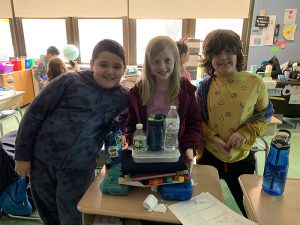 Three third grade students stand at a desk that is holding two pencil caseswith a piece of paper on it. On top of the paper are lots of things, including a cup and water bottle.
