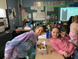 Two girls smile as they lean on a desk. There is a piece of paper across books and on the paper are blocks.