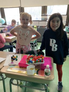 Two third-grade girls stand by a desk. They are showing a piece of paper across two pencil cases and on top of the paper are dozens of multi-colored blocks.