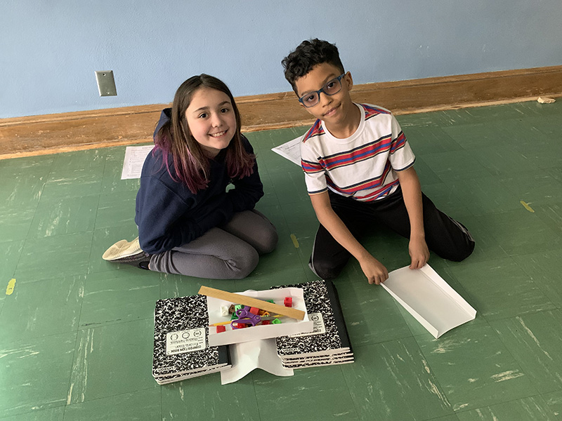 Two third-grade students sit on the floor. They have two stacks of three notebooks on the floor with a piece of paper across te top and one folded underneath the other. There are blocks and a ruler on top of the paper.