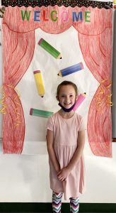 A girl wearing apink dress stands in front of graphic that says welcome in all different colors. She is smiling