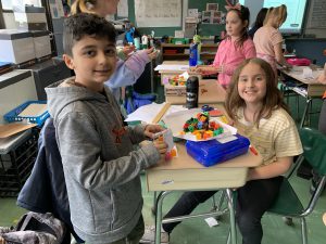 A boy and a girl in a classroom by a desk. There is paper on top of two plastic pencil cases and on top of the paper are many multi-color blocks.