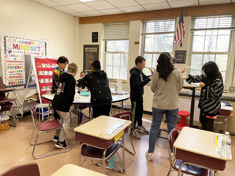 Two groups of three students each stand around tables working together. Beyond them are windows.