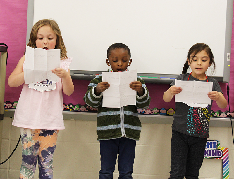 Three kindergarten kids stand reciting a poem. There are girls on either side and a boy in the center. 