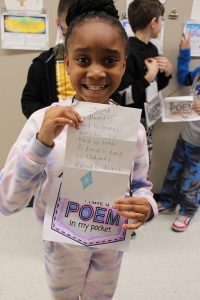 A girl with her dark hair in a bun smiles holding a piece of paper with a poem on it. It is  coming out of a pouch that says I have a Poem in my pocket