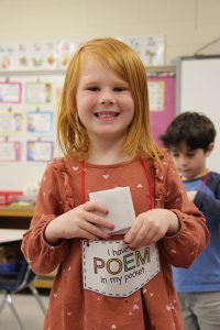 A little girl with shoulder-length reddish blonde hair smiles broadly as she puts a folder piece of paper in a pouch that says I have a Poem in my Pocket.