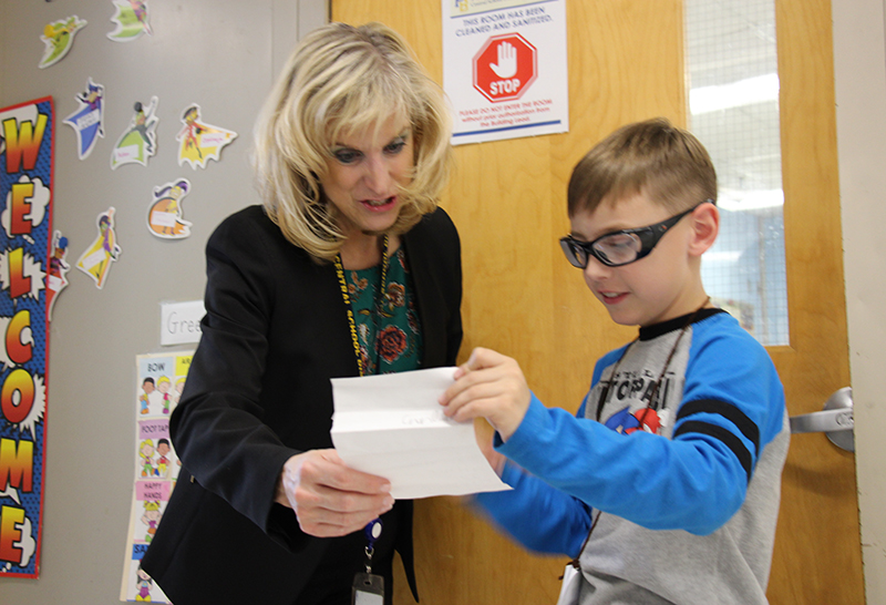 A woman with blonde hair, wearing a dark jacket stands with an elementary boy looking at a piece of paper he is showing her. He has short light hair and is wearing glasses, and a blue and gray long-sleeve shirt.