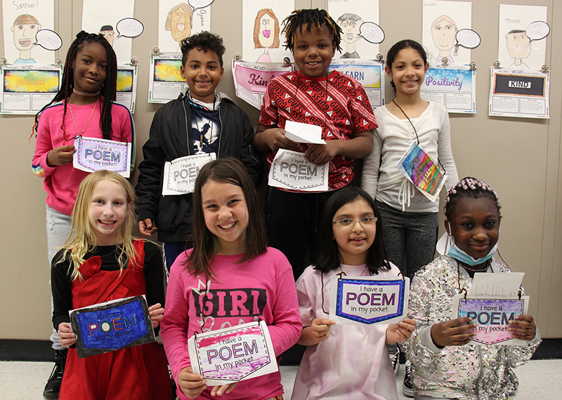 Eight third-graders stand in two rows of four, all holding paper that says Poem in my Pocket. They are smiling.