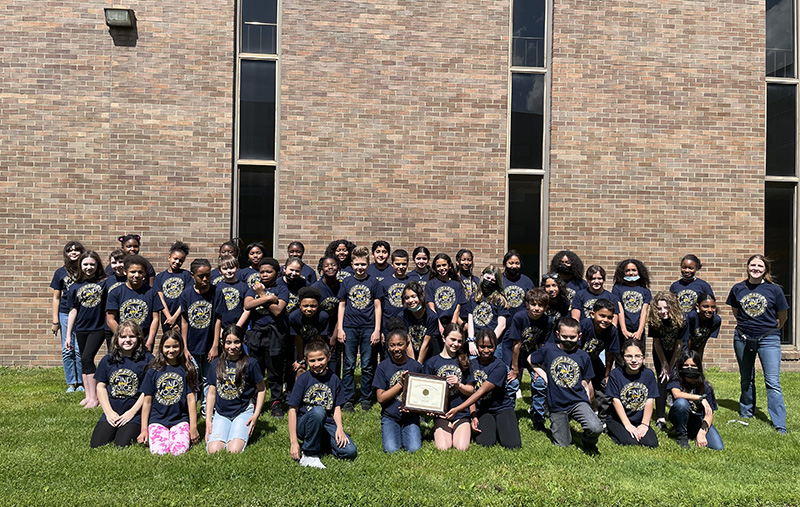 A large group of fifth-grade elementary students stand outside on green grass in front of a brick building. They are all wearing dark tshirts.