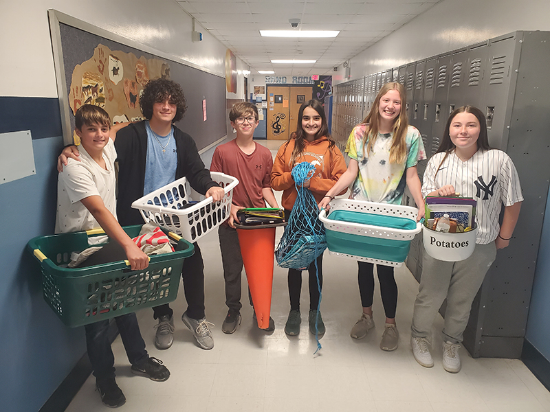 A group of six middle school students stand together smiling. Three boys on the left and three girls on the right. They are carrying their school supplies in laundry baskets, a mesh hamper, in a traffic cone and a white pot that says potatoes.