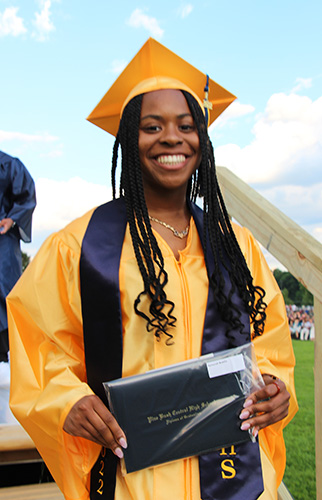 A young woman in a gold cap and gown with blue sash around her shoulders is smiling broadly. She has long dark braids.