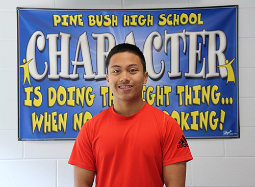 A young man smiles. He has short dark hair and is wearing an orange tshirt. Behind him is a sign that says Pine Bush High School - Character is doing the right thing when no one is looking.