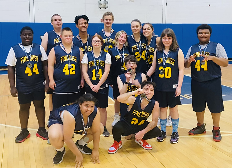 A group of 15 high school students wearing navy blue basketball uniforms with gold numbers on them. They say Pine Bush. The players have medals around their necks.
