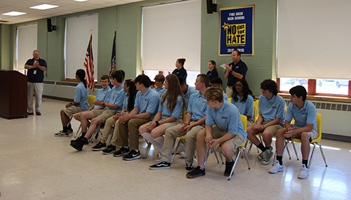 A man stands at a podium on the left and speaks to two rows of high school students, all wearing light blue polo shirts and tan pants.