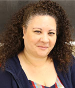 A woman with long curly hair, wearing a striped shirt and dark blue sweater smiles.