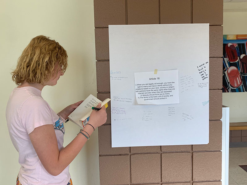 A young woman with short blonde hair holds a book and looks at a poster.