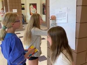 Three students stand in front of a pillar that has a poster on it. One girl with long blonde hair writes on the poster while the other two wait their turn.