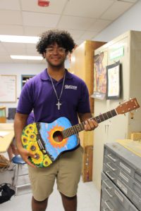 A young man with curly dark hair, wearing glasses, smiles. He is wearing a purple short sleeve shirt and holds a guitar that is painted with sunflowers on the bottom and blue above.