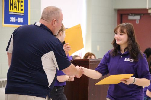 A man in a blue and white polo shirt shakes hands with a young woman, with shoulder-length brown hair and wearing a purple polo. She is holding an envelope in her hand.