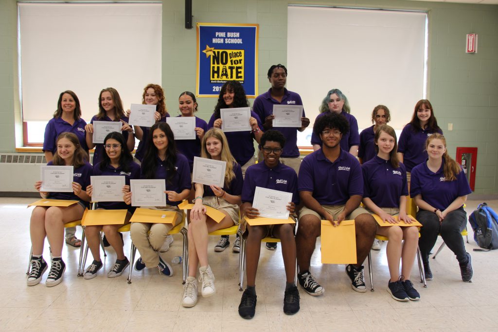 A large group of high school kids, all wearing purple polo shirts and khaki pants, are holding certificates. One row is sitting in front and another is standing behind.