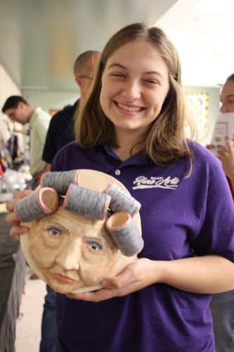 A young woman with shoulder-length light hair, wearing a purple polo shirt smiles. She is holding a ceramic piece of art she made - a woman with gray hair in large rollers.