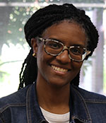 A woman in a white shirt with a denim jacket on, smiles. She is wearing glasses and her hair is braided and pulled back.