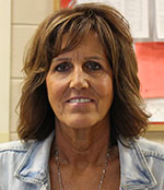 A woman with shoulder length brown hair, wearing a light blue denim jacket. She is smiling.