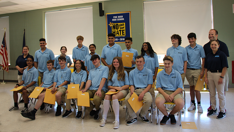 A group of about 15 high school students all wearing light blue shirts and tan pants. Half sit in chairs in front, and the other half stand behind them. There is also a man standing on the right side next to the students.