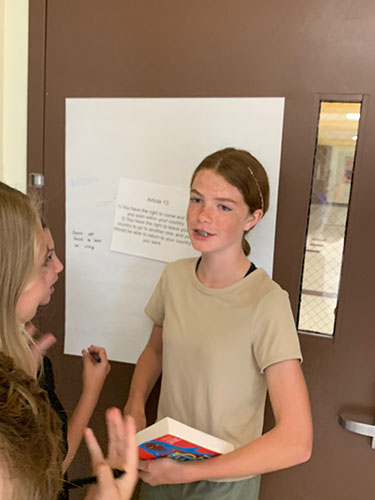 Three young women stand in front of a door with a poster on it. One girl with her dark hair pulled back in a ponytail . wo girls are talking to her.