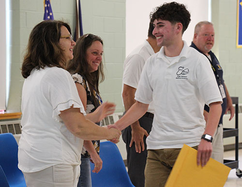 A young man with short curly dark hair smiles and shakes the hand of a woman who is smiling back. He is wearing a white shirt and holding a large manila envelope.