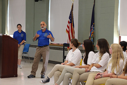 A man stands a few feet away from a dais and is talking to a group of high school students who are seated and all wearing white shirts and tan pants. Behind him is a woman also listening.