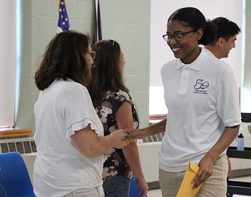 A high school girl wearing a white shirt and glasses, has her dark hair pulled back and shakes the hand of a woman with shoulder-length dark hair. 