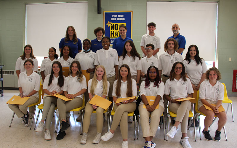 A group of 18 high school students smile and hold their manila envelopes in their hands. Half are sitting in seats in the front row, others are standing behind them. There is a third row with one student and four adults. The students are all wearing white shirts and tan pants. The adults have blue shirts on.