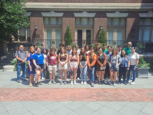 A large group of high school students and a few adults stand outside on a sunny day in front of a brick building.