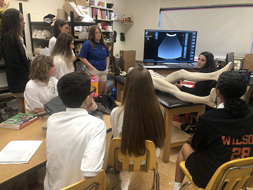 A group of seven high school students stand around a mannequin that is lying on a bed. An adult is performing a simulated sonogram and the results are on a monitor above them.