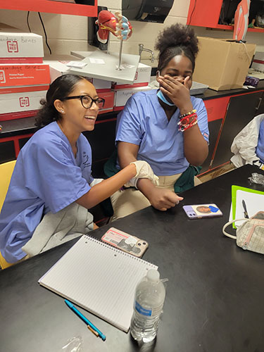 Two high school girls dressed in blue scrubs sit together. The girl on the left is smiling, wearing glasses and putting a bandage on the other girl's wrist. She is laughing, puttin gher hand over her mouth.