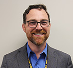 A man with short dark hair, close beard and mustache, wearing glasses smiles. He has a dark blue collared shirt and gray jacket on. 